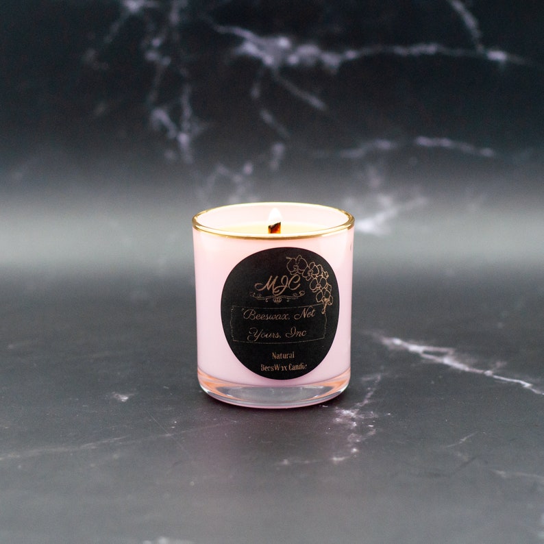 May include: A light pink candle in a glass jar with a gold rim. A black circular label features the text MJC, Beeswax, Not Yours, Inc, and Natural BeesWax Candle. The candle has a single lit wick, and the wax inside is a pale cream color.