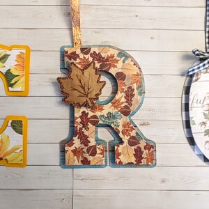 May include: Decorative wooden letters 'G', 'R', and 'C' with fall and floral themes. The 'G' features sunflowers, the 'R' has autumn leaves, and the 'C' displays pumpkins and cotton. Each letter is adorned with a ribbon for hanging.