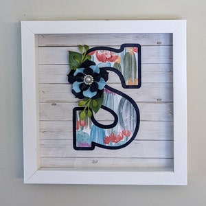 May include: A white framed shadow box with a black outlined letter "S" decorated with a blue and black flower and a cactus print background.