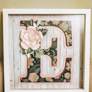 May include: A framed wall decor featuring a large pink letter E with a floral design. The letter is layered with a floral pattern and a pink border.
