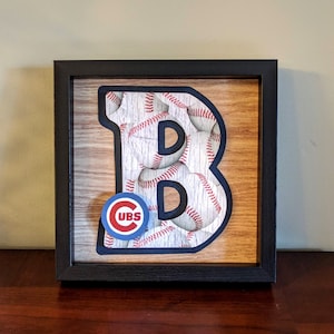 May include: A framed wooden sign with a black frame. The sign features a large letter "B" made of baseballs. The letter "B" is surrounded by a wood grain background. A small circular sticker with the words "CUBS" in red letters on a blue background is in the lower left corner of the sign.