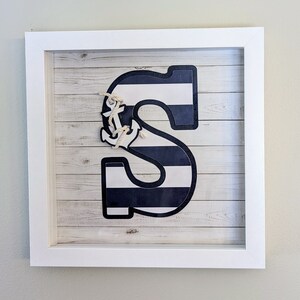 May include: A white framed picture with a nautical theme. The picture features a large blue and white striped letter "S" with a small white anchor on top. The background is a woodgrain pattern.
