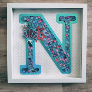 May include: A white framed picture with a turquoise letter N decorated with mermaid scales, a pink seashell, and red coral.