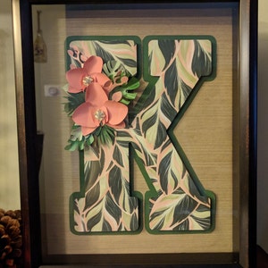 May include: A framed letter "K" with a green and pink tropical leaf pattern. The letter is decorated with pink paper flowers.