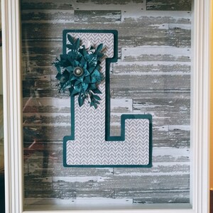 May include: A white frame with a teal letter L decorated with a teal paper flower and green leaves. The background is a woodgrain pattern.