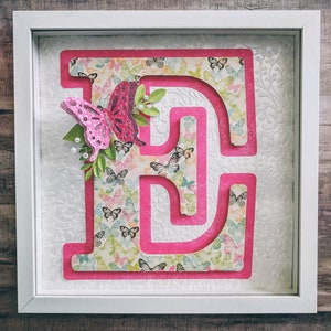 May include: A white frame with a pink letter E cutout. The letter is layered on top of a patterned paper with a butterfly design. The butterfly is pink and has glitter accents.