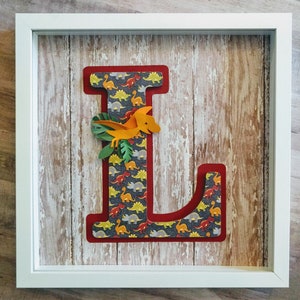 May include: A white frame with a red and white letter L decorated with a dinosaur pattern. A small orange dinosaur with green leaves is attached to the letter.