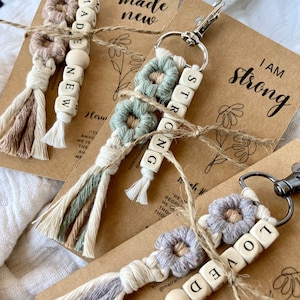 May include: Three keychains with colourful macrame tassels and wooden beads spelling out the words "Made New", "Strong", and "Loved". Each keychain is attached to a silver key ring and comes with a brown card with the words "I am made new", "I am strong", and "I am loved" written on them.