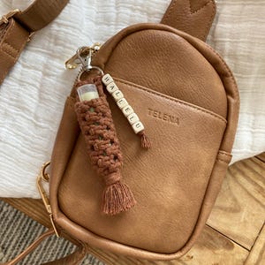 May include: A brown leather crossbody bag with a zip fastening and a gold metal buckle. The bag has a small pocket on the front with the word "TELENA" embossed on it. A keychain with the word "HALLELU" is attached to the bag.