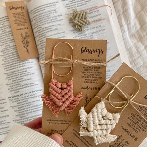 Peut inclure: Deux marque-pages en macramé faits à la main avec un clip doré et rose doré. Les marque-pages sont en forme de cœur et de colombe. Les marque-pages sont sur un fond brun avec le texte "blessings" et un verset biblique.
