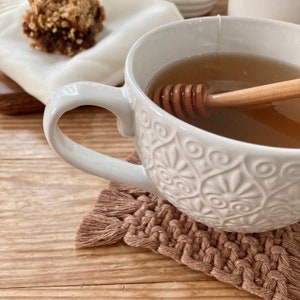 May include: A white ceramic mug with an intricate floral design, filled with a light brown liquid and a wooden honey dipper. The mug is sitting on a brown woven coaster.
