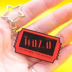 May include: Clear acrylic keychain with a red and black ticket design. The ticket reads "THE WAYLAY" with the text "ONE FREE DRINK ENJOY YOUR STAY!" A silver star-shaped clasp is attached to the keychain.