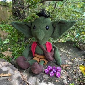 May include: A green plush goblin toy with brown boots, a red waistcoat, and a brown bag. The goblin is sitting on a rock with purple dice scattered around it.
