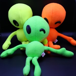 May include: Three plush alien toys in shades of green, orange, and yellow. Each has a round head, black oval eyes, and small arms and legs. One green alien is winking. The toys are soft and cuddly.