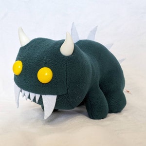 Könnte beinhalten: Ein grünes Plüschmonster mit weißen Hörnern und Zähnen, gelben Augen und weißen Stacheln auf dem Rücken.