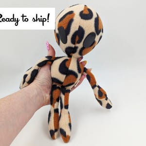 May include: A plush toy in the shape of a humanoid with a leopard print pattern. The toy has black eyes and brown and black fur.