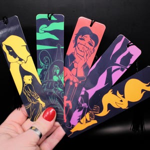 May include: Five rectangular bookmarks with black tassels, each featuring distinct graphic illustrations in vibrant colours including yellow, green, coral, pink, and orange against dark backgrounds, showcasing various character and nature-themed designs.