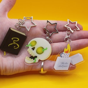 May include: A selection of acrylic keyrings featuring a skull with yellow eyes, a book with gold detailing, and a lighter with the text "This too shall pass." Each keyring has a silver star-shaped clasp.