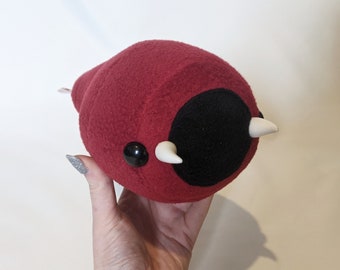 May include: Red plush toy with a black circle and two white horns. The toy is made of soft, fuzzy fabric.