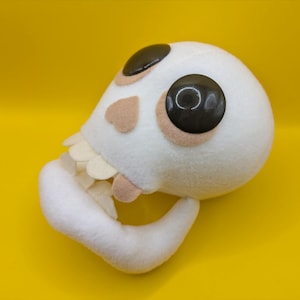 May include: A plush, white skull-shaped toy with large, dark brown eyes and tan accents. The skull has a curved jawline with off-white teeth. The toy is set against a bright yellow background.
