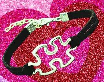 May include: A black leather bracelet with a silver puzzle piece charm. The bracelet is on a pink and purple glitter background.