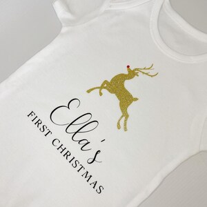 Puede incluir: Body de beb&eacute; blanco con el texto "Ella's First Christmas" en escritura negra. Un reno dorado con purpurina y nariz roja aparece en la parte delantera. El body es una prenda de manga corta.