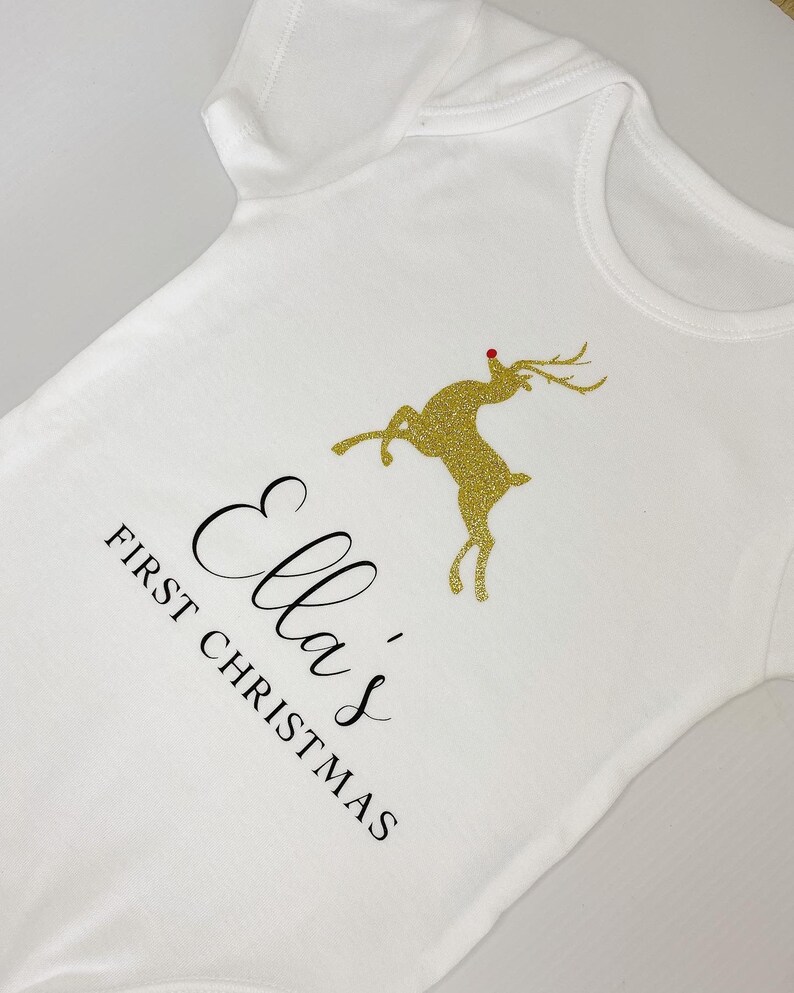 Puede incluir: Body de beb&eacute; blanco con el texto "Ella's First Christmas" en escritura negra. Un reno dorado con purpurina y nariz roja aparece en la parte delantera. El body es una prenda de manga corta.