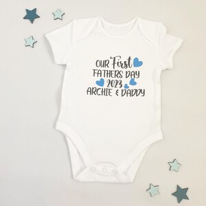 May include: White baby onesie with blue hearts and the text "Our First Father's Day 2023 Archie & Daddy"