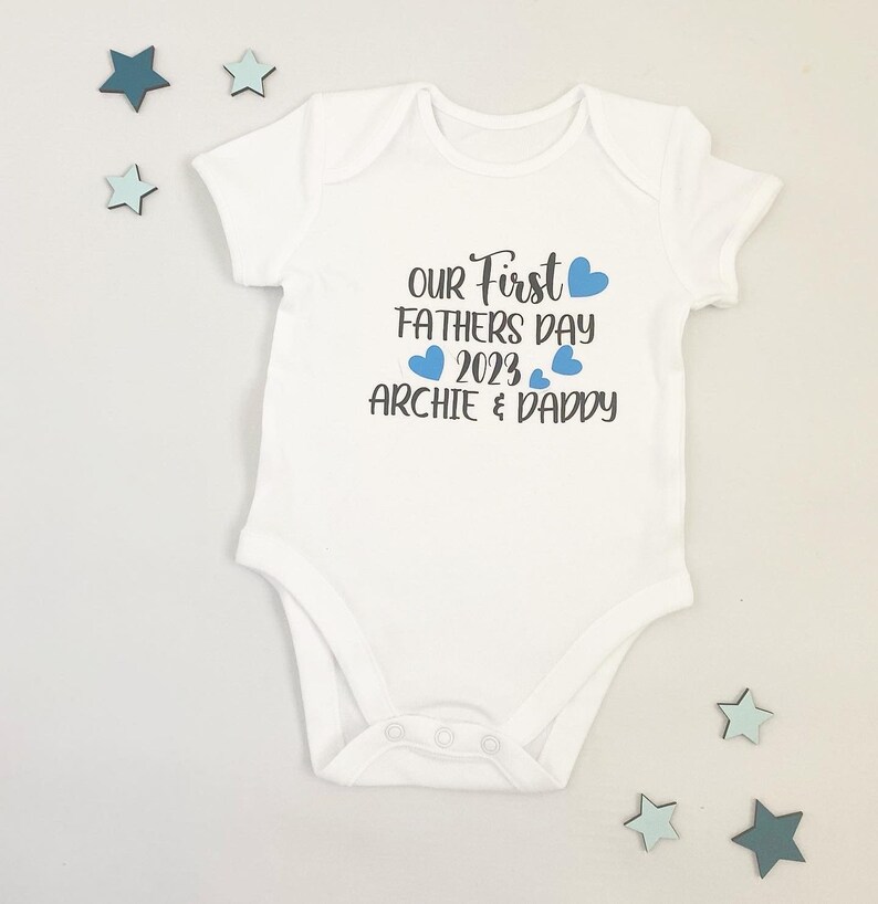 May include: White baby onesie with blue hearts and the text "Our First Father's Day 2023 Archie & Daddy"