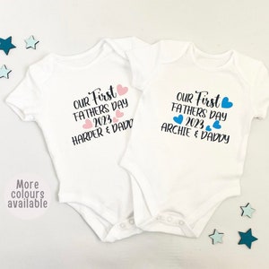 May include: Two white baby onesies with short sleeves. The onesies have the text "Our First Father's Day 2023" with pink and blue hearts. One onesie says "Harper & Daddy" and the other says "Archie & Daddy".