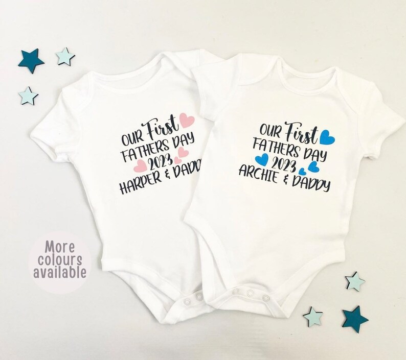 May include: Two white baby onesies with short sleeves. The onesies have the text "Our First Father's Day 2023" with pink and blue hearts. One onesie says "Harper & Daddy" and the other says "Archie & Daddy".