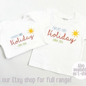 May include: Two white short-sleeved shirts with graphic prints. One features the text "ISHAQ'S FIRST Holiday" in blue and red with "CORFU 2024" in green and a yellow sun. The other says "Layla's FIRST Holiday" in blue and red with "SPAIN 2024" in green.