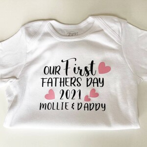 May include: White baby onesie with black text that reads "Our First Father's Day 2021 Mollie & Daddy" with pink hearts.
