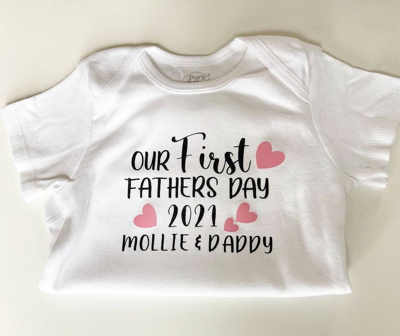 May include: White baby onesie with black text that reads "Our First Father's Day 2021 Mollie & Daddy" with pink hearts.