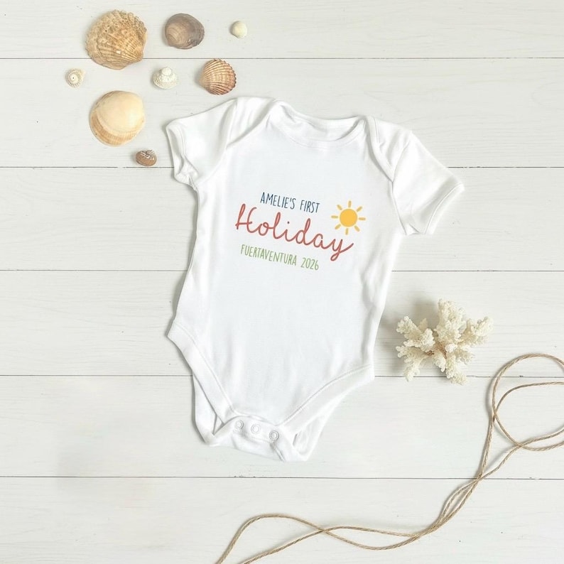 May include: A white short-sleeved baby bodysuit featuring the text AMELIE'S FIRST Holiday FUERTEVENTURA 2026 in blue, red, and green, accompanied by a yellow sun graphic. The garment is surrounded by various seashells and a piece of white coral.