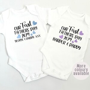 May include: Two white baby onesies with the text "Our First Father's Day 2024" and the names "George & Daddy" and "Harper & Daddy" printed on them. The onesies have blue and purple hearts printed on them.