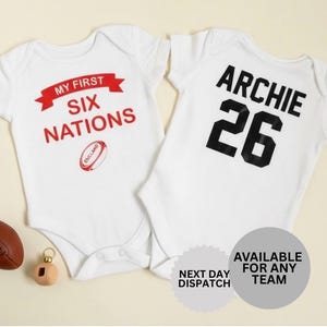 Könnte beinhalten: Zwei weiße Baby-Bodys. Einer hat den roten Aufdruck "MY FIRST SIX NATIONS" mit einem Rugbyball-Motiv. Der andere hat "ARCHIE 26" in Schwarz. Ein brauner Rugbyball und ein Holzspielzeug sind ebenfalls vorhanden.
