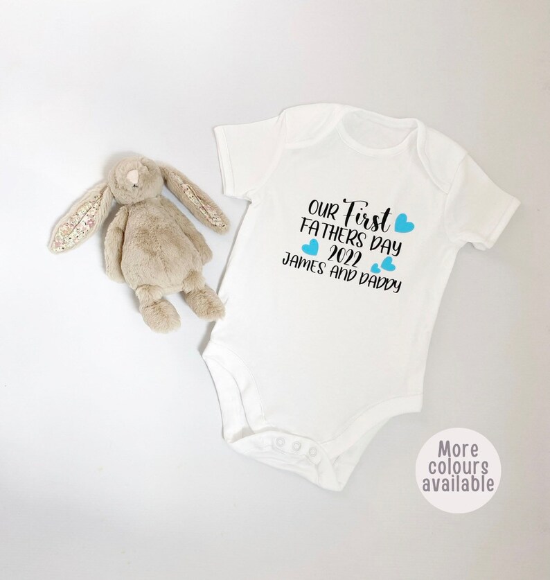 May include: White baby onesie with the text "Our First Father's Day 2022 James and Daddy" in black with blue hearts.