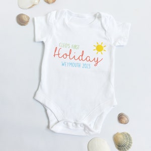 May include: A white short-sleeved baby bodysuit featuring the printed text Cleo's First Holiday in green and red, the word Weymouth 2023 in blue, and a yellow sun graphic. The garment has a round neckline and three snap closures at the bottom.