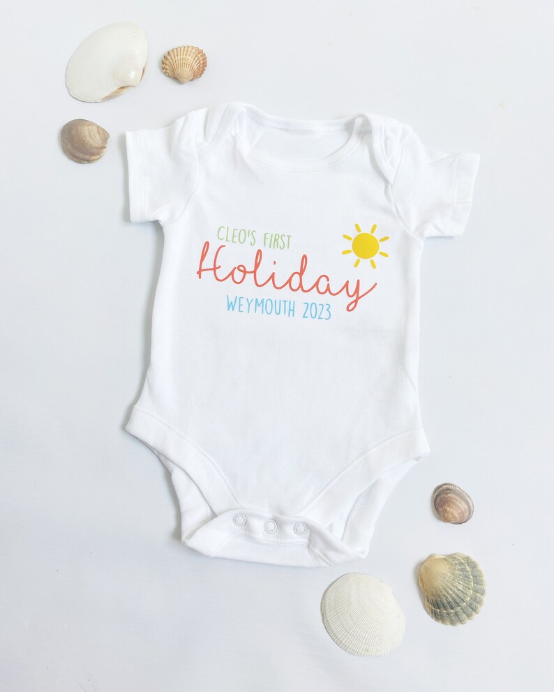 May include: A white short-sleeved baby bodysuit featuring the printed text Cleo's First Holiday in green and red, the word Weymouth 2023 in blue, and a yellow sun graphic. The garment has a round neckline and three snap closures at the bottom.