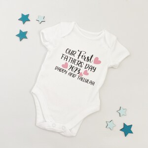 May include: White baby onesie with the text "Our First Fathers Day 2023 Daddy and Tallulah" in black with pink hearts.
