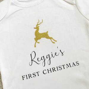 Puede incluir: Body de beb&eacute; blanco con un dise&ntilde;o de reno dorado con purpurina y el texto "Reggie's FIRST CHRISTMAS" en escritura negra. El reno tiene una nariz roja. Una prenda festiva para la primera Navidad de un beb&eacute;.