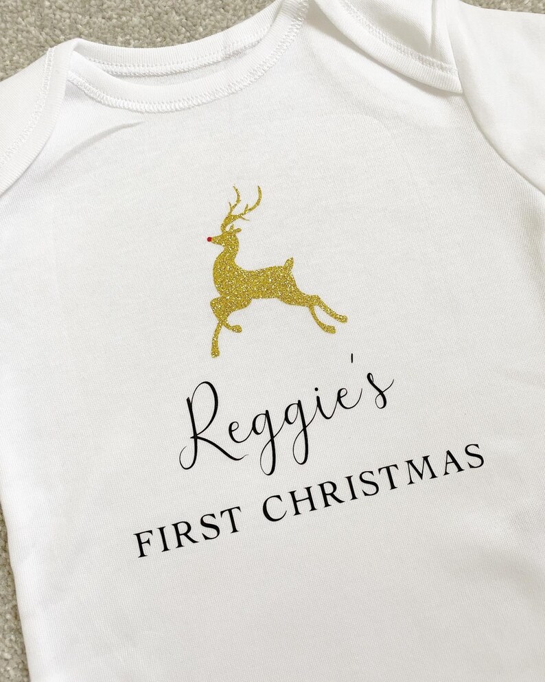 Puede incluir: Body de beb&eacute; blanco con un dise&ntilde;o de reno dorado con purpurina y el texto "Reggie's FIRST CHRISTMAS" en escritura negra. El reno tiene una nariz roja. Una prenda festiva para la primera Navidad de un beb&eacute;.