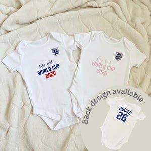 May include: Two white short-sleeved baby bodysuits featuring the England national team crest and the text My first WORLD CUP 2026. One bodysuit has blue and red text, the other has light pink text. A smaller inset shows a back design with OSCAR 26.