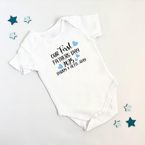 May include: White baby onesie with the text "Our First Father's Day 2025 Daddy & Alfie-Ray" in black lettering and blue hearts.