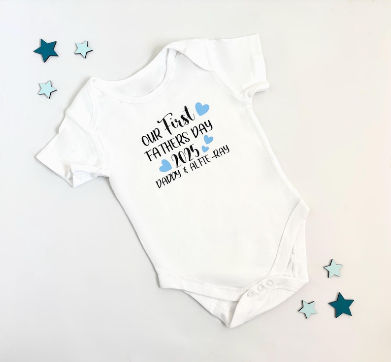 May include: White baby onesie with the text "Our First Father's Day 2025 Daddy & Alfie-Ray" in black lettering and blue hearts.