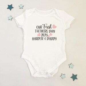 May include: White baby onesie with the text "Our First Fathers Day 2023 Harper & Daddy" in black with pink hearts.