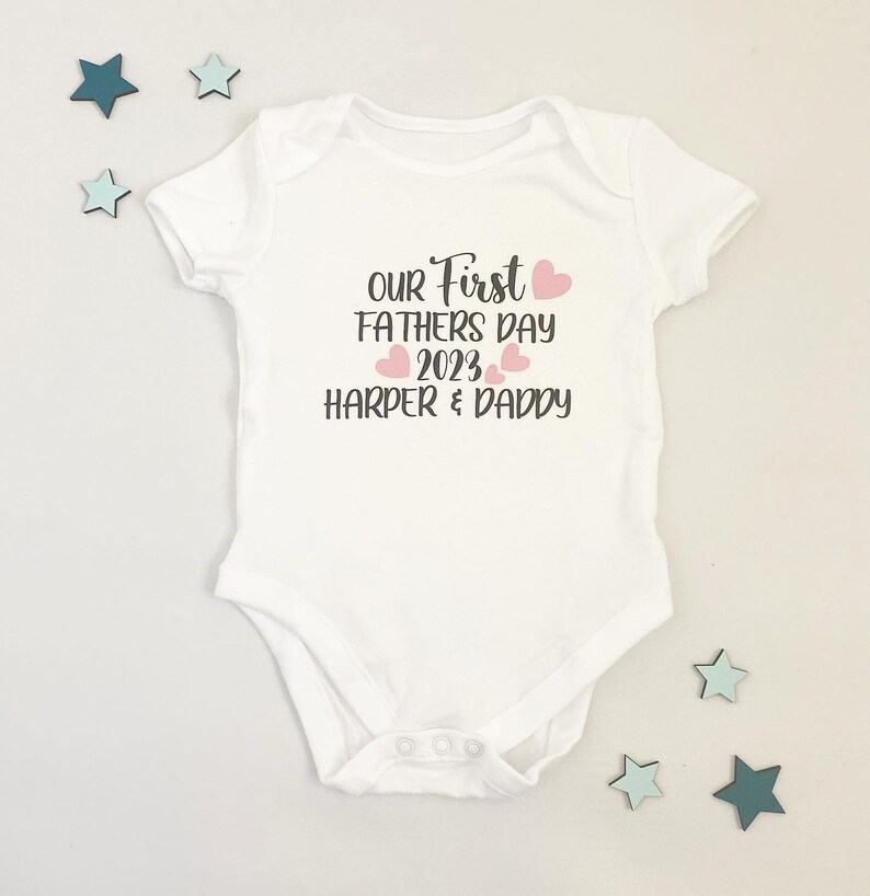 May include: White baby onesie with the text "Our First Fathers Day 2023 Harper & Daddy" in black with pink hearts.