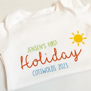 May include: A white short-sleeved baby bodysuit featuring the green text JENSEN'S FIRST, the orange script text Holiday, the blue text COTSWOLDS 2023, and a yellow sun graphic with rays positioned to the right of the text.
