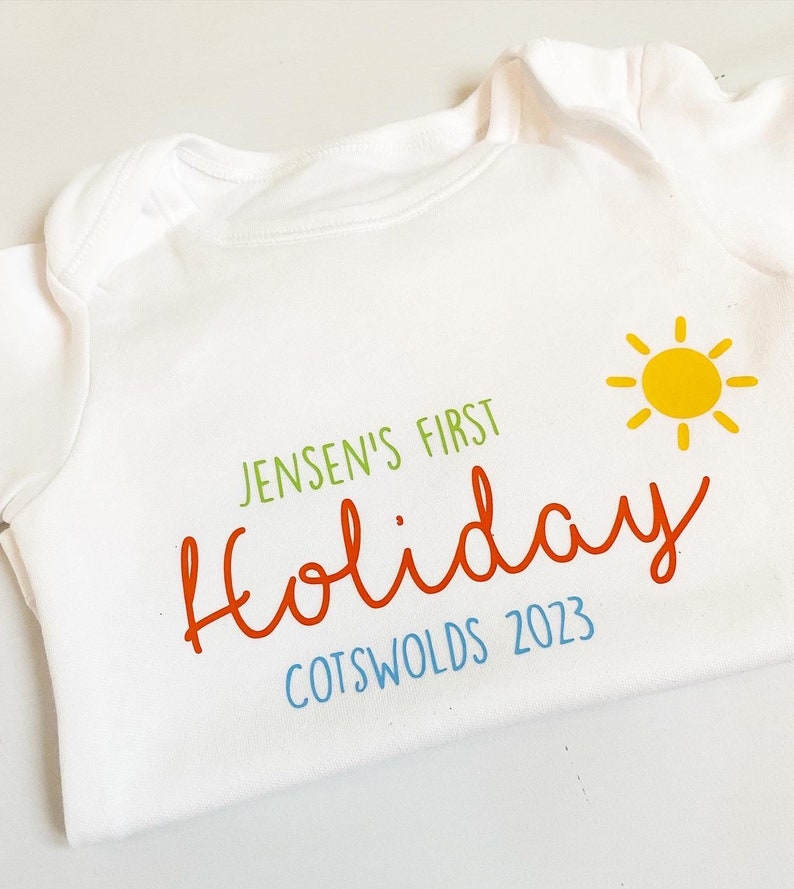 May include: A white short-sleeved baby bodysuit featuring the green text JENSEN'S FIRST, the orange script text Holiday, the blue text COTSWOLDS 2023, and a yellow sun graphic with rays positioned to the right of the text.